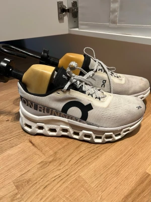 Vita On Running sneakers med snörning - Snygga vita sneakers från On Running med svart logga på sidan och cool, chunky sula. Skorna har svart och vit plös, samt vita snören med svarta detaljer. Ovandelen är i mesh-liknande syntetmaterial som andas. Perfekta för dig som gillar sportig och modern stil. Observer att det är ett litet hål som är sytt på ena foten, se bild. Storlek 40, men jag upplever de som lite små. 