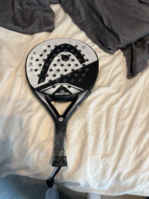 Svart och vit padelracket Head Master - Padelracket från Head i svart och vitt med tydlig logga och texten 'Master' på framsidan. Racketen har rund form, perforerad yta och grepp med plastskydd. Perfekt för dig som gillar padel och vill ha en snygg och funktionell racket.