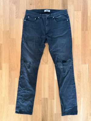 Svarta jeans med slitningar från Acne - Snygga svarta jeans från Acne med raka ben och slitna detaljer på knäna. Klassisk femficksmodell med silverfärgade nitar och knapp. Jeansen har en modern, avslappnad look och är tillverkade i ett mjukt denimtyg. Storlek 34/32. 