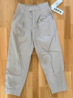 Beige chinos från Hope - Snygga beige chinos från Hope med avslappnad passform och hög midja. Byxorna har raka ben, bälteshällor och diskreta sidofickor. Tillverkade i ett mjukt bomullsmaterial som känns skönt mot huden. Perfekta för en clean och stilren look.