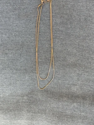 Tunt guldigt halsband i dubbel kedja - Säljer ett stilrent halsband med två tunna, guldiga kedjor som ligger i lager. Halsbandet har en enkel och modern design med små detaljer vid låset. Perfekt för dig som gillar minimalistiska smycken och vill ha något som sticker ut utan att vara för mycket.