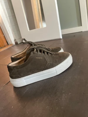 Bruna sneakers från Axel Arigato -  Nypris 2000:- Snygga bruna sneakers från Axel Arigato i mocka med vit platåsula. Klassisk låg modell med snörning och diskret logga på sidan. Perfekta för en stilren och trendig look till jeans eller andra vardagsoutfits. Inte skärsilt slitna syntesen på användning är bara invändigt bud tas