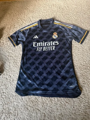 Real Madrid bortatröja Adidas M - Snygg Real Madrid bortatröja från Adidas i mörkblått med mönstrad design. Tröjan har gula och vita detaljer på axlarna, klubbmärke på bröstet och 'Emirates Fly Better' tryckt framtill. Tillverkad i lätt och ventilerande material, perfekt för fotbollsträning. Aldrig använd tidigare pris kan diskuteras!