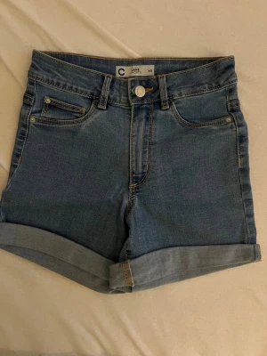 Blå jeansshorts från C Jane - Snygga blå jeansshorts från C Jane med klassisk femficksdesign och uppvikta benslut. Perfekta för varma dagar och enkla att matcha med allt. Stängs med knapp och dragkedja. Tidlös modell som alltid funkar.
