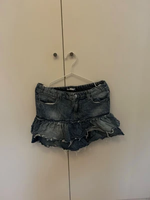 Blå jeanskjol med volang och slitningar - Säljer en blå jeanskjol med volangkant och slitna detaljer. Kjolen har fickor fram och bak, samt råa kanter för en trendig look. Perfekt för dig som gillar en avslappnad och cool stil.