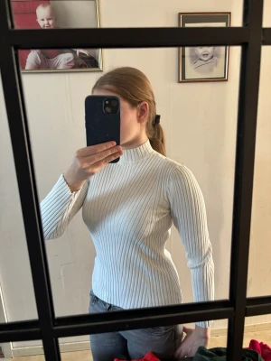 Vit ribbad polotröja - Säljer en vit ribbad polotröja med lång ärm och figurnära passform. Tröjan har hög hals och är tillverkad i ett stretchigt material som sitter snyggt på kroppen. Perfekt att styla med jeans eller kjol för en clean look. Lite missfärgad på en liten dela av polokragen där de finns en gammal sminkfläck som knappt syns.