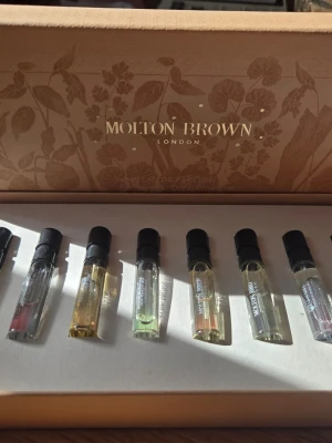Molton Brown parfymset med 8 dofter - Upptäck ett exklusivt parfymset från Molton Brown med åtta olika doftprover i små glasflaskor. Setet levereras i en elegant beige ask med blommönster och guldtext. Perfekt för dig som vill testa flera lyxiga dofter från London-märket.