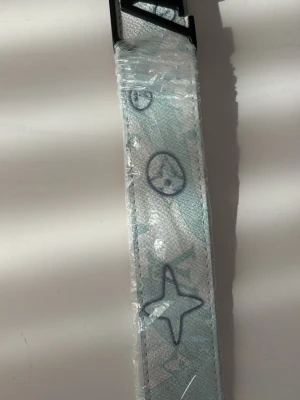 Turkost bälte med stjärnmönster - Snyggt vitt bälte med ljusblått och blått stjärnmönster samt cirklar. Bältet har en stilren design och ett spänne i svart. Perfekt för att lyfta din outfit med en unik detalj. Skriv om ni har frpgor eller vill se fler bilder