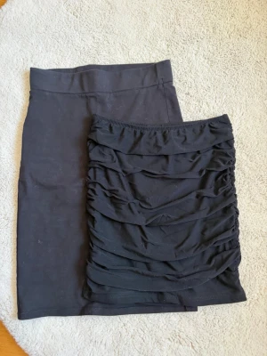 Svart tight kjol med rynkade detaljer - Säljer två svarta tighta kjolar med snygga rynkade detaljer framtill. Kjolen är kort och har en kroppsnära passform som framhäver figuren. Perfekt för dig som gillar en enkel men ändå trendig look. Materialet känns mjukt och stretchigt.