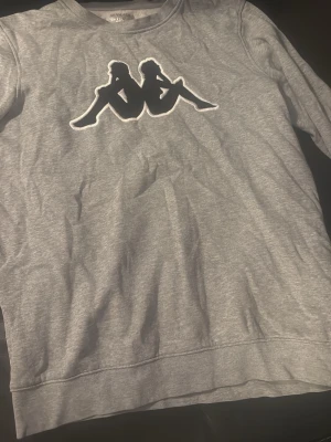 Grå sweatshirt från Kappa - Säljer en grå sweatshirt från Kappa med det klassiska logotrycket framtill. Tröjan är i mjukt material och har en avslappnad passform, perfekt för en chill och sportig stil. Långärmad och utan huva.