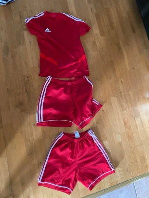 Rött Adidas träningsset med shorts - Säljer ett rött träningsset från Adidas med en t-shirt och två par matchande shorts. Alla delar har vita ränder längs sidorna och Adidas-logga på tröjan. Perfekt för fotboll eller annan träning.