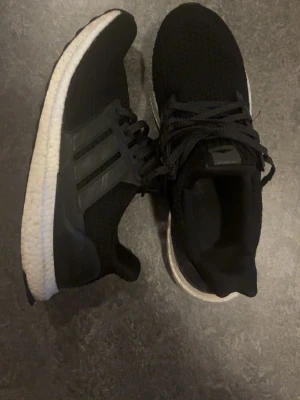 Svarta Adidas Ultraboost sneakers - Säljer ett par svarta Adidas Ultraboost sneakers med klassiska tre ränder på sidan. Ovandelen är i stickad syntet och sulan är vit och riktigt bekväm. Perfekta för dig som vill ha både stil och komfort i vardagen.