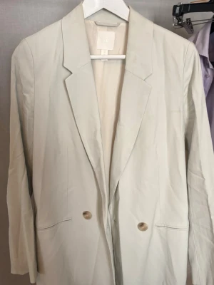 Beige oversized kavaj från H&M - Stilren oversized kavaj i beige från H&M med klassisk krage och två knappar framtill. Långa ärmar och två fickor framtill. Perfekt för en trendig och avslappnad look.