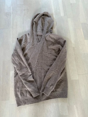 Beige stickad hoodie med v-ringning - Mysig beige stickad hoodie med v-ringning och långa ärmar. Tröjan har ribbade muddar vid ärmslut och nederkant. Perfekt för en avslappnad och stilren look.