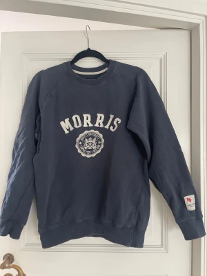 Blå sweatshirt från Morris - Mysig blå sweatshirt från Morris med vit logga och emblem framtill. Tröjan har rund halsringning, långa ärmar och ribbade muddar vid ärmslut och nederkant. Perfekt för en avslappnad och stilren look. Denna är vintage och riktigt snygg för både killar och tjejer.