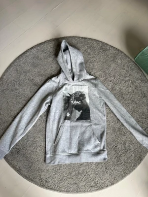 Grå hoodie med Tupac-tryck - Snygg grå hoodie med stor svartvit Tupac-bild på bröstet och texten 'TUPAC'. Klassisk känguruficka framtill och huva. Perfekt för dig som gillar streetwear och vill ha en bekväm och stilren look.