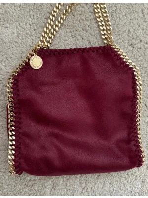Burgundy Stella McCartney handväska - Snygg vinröd handväska från Stella McCartney med guldfärgade kedjor. Har haft den i 2 år men den har tyvärr inte kommit så mycket till användning. Har alla äkthetsbevis och original tygpåse som självklart medföljer❤️