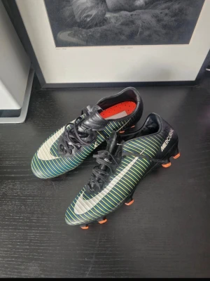 Nike Mercurial fotbollsskor svart/grön - Säljer ett par Nike Mercurial fotbollsskor med svart bas, gröna och vita ränder samt orange dobbar. Skorna har en klassisk låg profil, snörning och syntetmaterial för bra bollkontroll. Perfekta för dig som vill ha stilrena och snabba skor på planen.