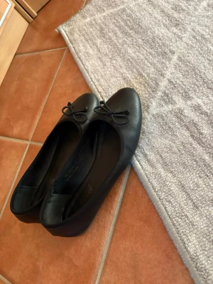 Svarta ballerinaskor med rosett - Säljer ett par klassiska svarta ballerinaskor med rund tå och liten rosett framtill. Skorna är i ett slätt, lätt glansigt material och har en enkel, stilren design som passar till det mesta. Perfekta för dig som gillar en tidlös look. Se defekt sista bild.