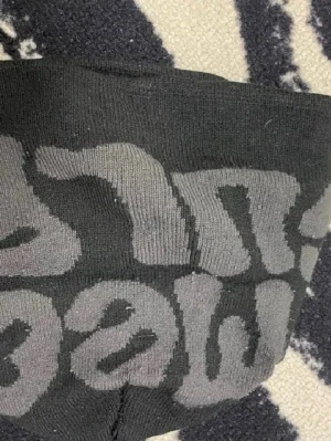 Svart mössa med grå text - Säljer en svart mössa med stor grå text i grafisk stil runt om. Mössan är stickad i mjukt material och har en klassisk, rundad form som sitter skönt på huvudet. Perfekt för dig som gillar streetwear och vill ha en enkel men snygg accessoar.