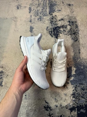 Adidas Ultra Boost vita sneakers - Fräscha Adidas Ultra Boost sneakers i helvitt med stickad ovandel och klassiska tre ränder på sidan. Skorna har en bekväm Boost-sula och svart yttersula. Perfekta för dig som gillar stilrena och sportiga sneakers med modern känsla.