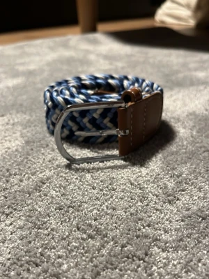 Blåvitt flätat bälte BVALDI - Snyggt flätat bälte i blått och vitt med brunt skinn vid spännet och silvrigt metallspänne. Perfekt accessoar för att lyfta din outfit med en sportig och fräsch känsla.