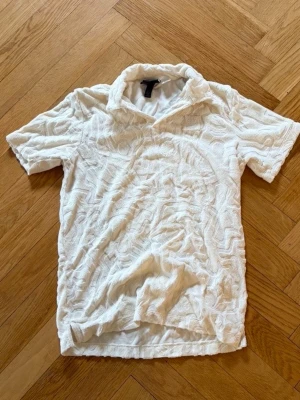 Vit frottéskjorta med krage - Säljer en vit skjorta från H&M i mjuk frotté med korta ärmar och klassisk krage. Skjortan har en unik struktur i tyget som ger en cool och avslappnad look. Perfekt för dig som gillar bekväma och trendiga plagg.