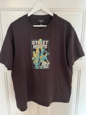 Svart Street Hoops t-shirt H&M - Cool svart t-shirt från H&M med färgglatt Street Hoops-tryck på bröstet. Motivet visar basketspelare i blått och gult med texten 'Street Hoops'. Klassisk passform och rund halsringning. Perfekt för en sportig och avslappnad stil.
