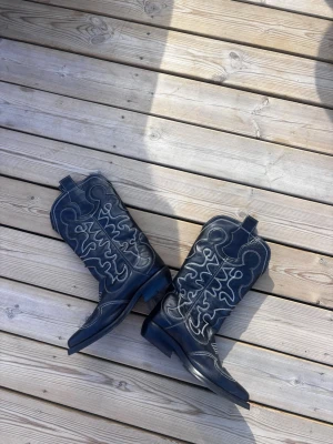 Svarta cowboy boots - Säljer ett par svarta cowboy boots i skinn med coola vita kontrastsömmar och klassiskt högt skaft. Bootsens spetsiga tå och dekorativa mönster ger en riktigt western vibe. Perfekta för festivaler och våren! Nypris ca 3000