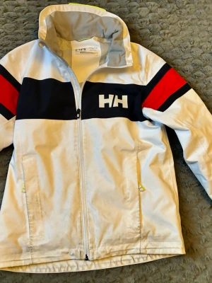 Vit Helly Hansen vindjacka med röd detalj - Snygg vit vindjacka från Helly Hansen med svarta och röda detaljer över bröst och ärmar. Jackan har dragkedja framtill, hög krage och HH-logga på bröstet. Perfekt för blåsiga dagar och sportig stil.