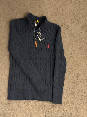 Mörkblå kabelstickad tröja Polo Ralph Lauren - Mörkblå kabelstickad tröja från Polo Ralph Lauren med halv dragkedja och ribbade muddar. Tröjan har en klassisk röd broderad logga på bröstet och är tillverkad i mjukt material. Perfekt för en stilren och avslappnad look.