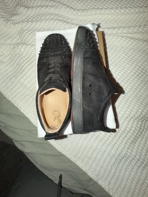 Svarta Louboutin sneakers med nitar - Svarta sneakers från Christian Louboutin med mattsvart ovandel i mocka och coola svarta nitar på tån. Skorna har den ikoniska röda sulan och svarta snören. Perfekta för dig som vill sticka ut med en edgy och lyxig stil. Skriv för mer frågor?