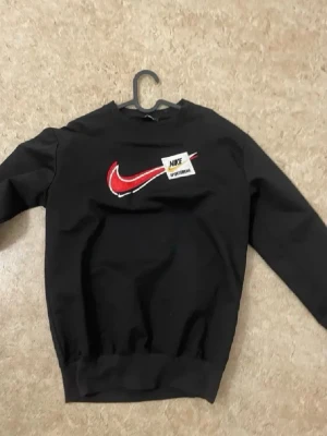 Svart sweatshirt från Nike - Cool svart sweatshirt från Nike med stor röd Swoosh-logga och en patch med texten 'Nike Sportswear' på bröstet. Tröjan har rund hals, långa ärmar och ribbade muddar. Perfekt för en sportig och avslappnad stil.