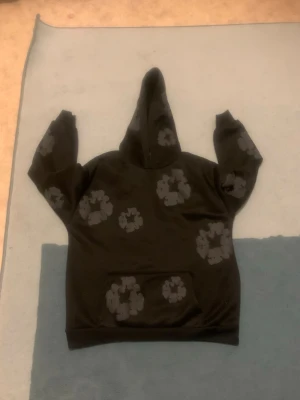 Svart hoodie med gråa blommor - Säljer en svart hoodie med gråa blommönster utspridda över hela tröjan. Hoodien har en klassisk huva och en stor magficka framtill. Perfekt för dig som gillar streetwear och vill sticka ut lite extra. Materialet känns mjukt och bekvämt.