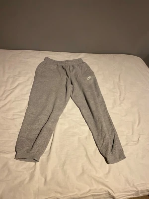Grå Nike mjukisbyxor med logga - Säljer ett par grå mjukisbyxor från Nike med vit logga på vänster sida. Byxorna har elastisk midja och muddar vid bensluten för en skön passform. Perfekt för chill dagar och sport. Materialet är mjukt och bekvämt, troligtvis bomullsblandning.