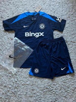 Chelsea träningsset från Nike [FAST PRIS] - Chelsea FC träningsset från Nike med tröja och shorts i marinblått och blått. Setet har klubbmärke, Nike-logga och BingX-tryck. Tillverkat i lätt och ventilerande polyester, perfekt för fotbollsträning. Snygg sportig look med korta ärmar och elastisk midja på shortsen.