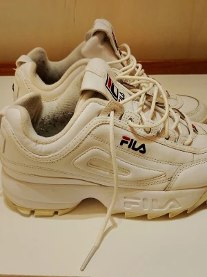 Vita Fila Disruptor sneakers - Chunky vita Fila Disruptor sneakers med kraftig sula och klassisk logga på sidan, plösen och hälen. Skorna har en retro vibe med detaljer i vitt och diskreta röda och blå inslag på loggan. Ovandelen är i syntetmaterial och textil, med snörning framtill.