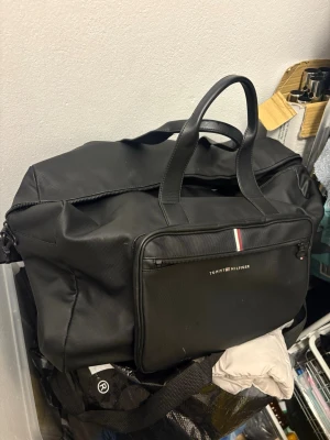 Svart weekendbag Tommy Hilfiger - Snygg svart weekendbag från Tommy Hilfiger med klassisk logga och röd-vit-blå detalj på framsidan. Väskan har både handtag och justerbar axelrem samt en stor ytterficka med dragkedja. Perfekt för resor eller gymmet.         Något litet vitt märke enligt bilderna - troligtvis från läckt tandkräm. 
