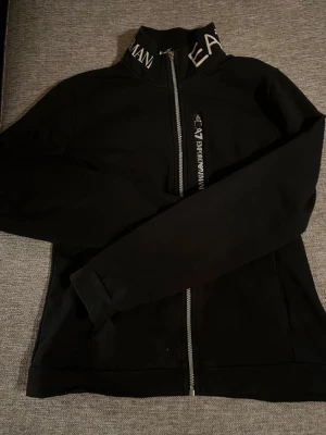 Svart set från Emporio Armani - Svart set från Emporio Armani EA7 med hel dragkedja och hög krage med vit logotyptext. Snygg och stilren design med diskreta logodetaljer på bröstet och nacken. Tillverkad i mjukt material som känns skönt mot huden. Perfekt för en trendig och sportig look. 