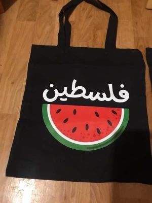 Svart tygkasse med vattenmelontryck - Cool svart shoppingväska i tyg med tryck av en stor vattenmelon och text på arabiska. Perfekt för att bära böcker, mat eller annat du behöver ha med dig. Väskan har långa handtag och är lätt att vika ihop när den inte används.