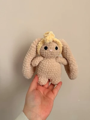 Beige virkad kanin med långa öron - Supergullig beige virkad kanin med långa öron, svarta ögon och liten rosa nos. Kaninen har en ljusgul virkad hårband och en vit svans. Mjuk och fluffig, perfekt som prydnad eller gosedjur.