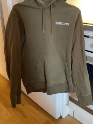 Brun hoodie från Helmut Lang - Brun hoodie från Helmut Lang med vit logga på bröstet. Klassisk känguruficka framtill, dragsko i huvan och ribbade muddar vid ärmslut och nederkant. Perfekt för en avslappnad och stilren look.