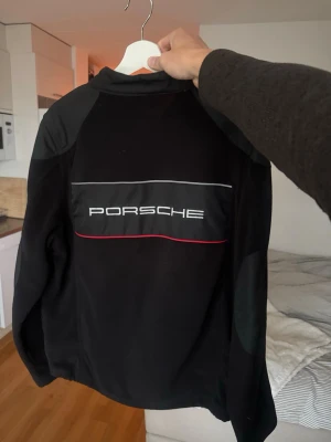 Svart Porsche jacka med dragkedja - Svart jacka från Porsche med hel dragkedja framtill och stora fickor. Jackan har diskreta grå detaljer och en vit Porsche-logga på ryggen med röd linje under. Perfekt för dig som gillar sportig och stilren design.