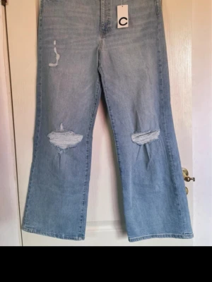 Ljusblåa bootcut jeans med slitningar - Ljusblåa jeans med bootcut-passform och slitna detaljer på båda knäna. Jeansen har hög midja och klassiska bakfickor. Perfekta för en avslappnad och trendig look.