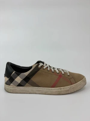Beige rutiga sneakers från Burberry - Snygga sneakers från Burberry i beige canvas med klassiskt rutmönster i svart, vitt och rött. Skorna har vita snören och svart hälparti i läder. Perfekta för dig som vill ha en stilren och trendig look med lyxig känsla.