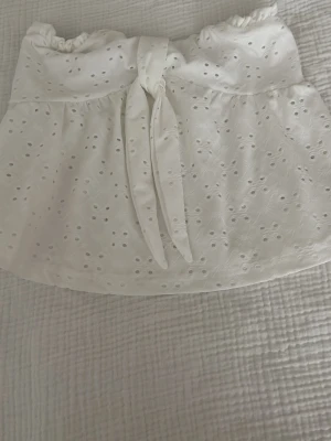 Vit broderad bandeau topp med knyt - Superfin vit bandeau topp med broderade hål och knyt framtill. Toppen är axelbandslös och har en söt, romantisk känsla med volangliknande fall. Perfekt för varma dagar och somriga outfits.