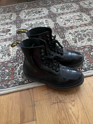 Svarta Dr. Martens lackstövletter - Säljer ett par klassiska svarta Dr. Martens stövletter i lackat läder. Modellen har hög snörning, grov sula och den ikoniska gula flärpen bak. Perfekta för dig som vill ha en edgy och tidlös look.
