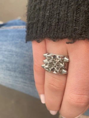 Silvrig ring från Edblad - Cool och chunky ring i silverfärgad metall med flera små konformade nitar på ovansidan. Ringen har en bred design och ger en edgy känsla till din stil. Perfekt för dig som gillar att sticka ut med dina accessoarer. Från Edblad 