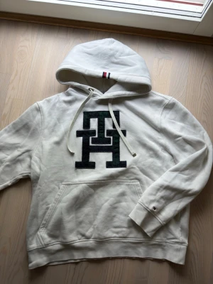 Vit hoodie från Tommy Hilfiger - Vit hoodie med stor TH-logga i grönt och marinblått på bröstet. Klassisk känguruficka och dragsko i huvan. Detalj med röd, vit och blå rand på huvan och liten logga på ärmslutet. Perfekt för en chill och stilren look.
