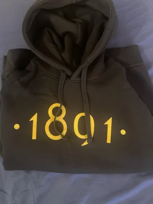 Svart AIK hoodie med gul text 1891 storlek S - Snygg svart hoodie med huva och dragsnören. På bröstet står det '1891' i stor gul text. Hoodien har en klassisk passform och magficka framtill. Perfekt för dig som gillar stilrena plagg med en twist. Bra skick inga defekter tyvärr för liten för mig.